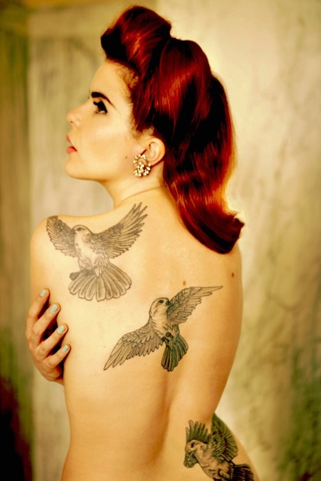 A woman with red hair styled in vintage waves poses with her back to the camera, showcasing three large bird tattoos—her "Out Without the Kids" moment. She wears ornate earrings set against a soft, yellow-toned background.