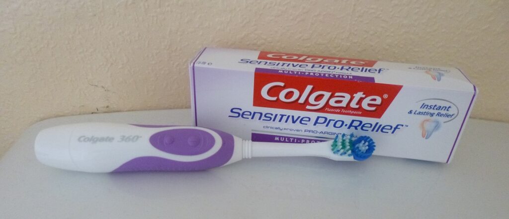 A Colgate 360° electric toothbrush with a purple handle sits in front of a box of Colgate Sensitive Pro-Relief toothpaste on a white surface against a beige wall.
