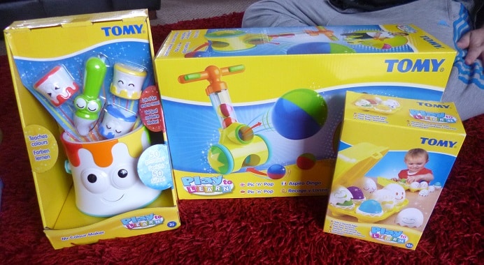 Three TOMY Play to Learn educational toys are on a red carpet: a colourful paint tin toy with brushes, a push-along ball toy, and a box with egg-shaped characters. Part of a person is visible in the background.