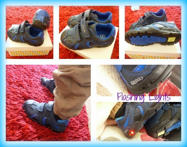 A pair of children's blue and black Clarks Velcro trainers is shown from multiple angles, including on a red carpet and worn by a child. One shoe sole displays a flashing red light. The words "Flashing Lights" appear on the image.