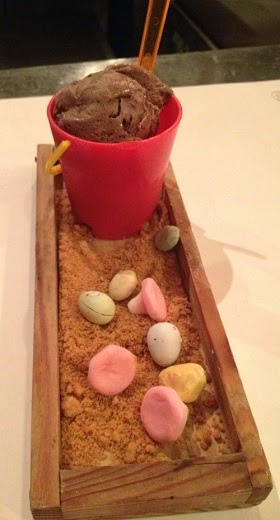 A dessert from The Botanist, served in a small red bucket with chocolate ice cream, sits on a tray of biscuit crumbs and colourful sweet stones—an edible beach scene perfect for a Family Meal at Trinity Leeds.