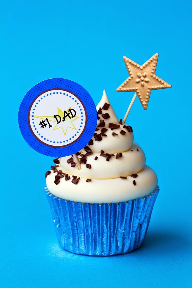 A cupcake with white icing and chocolate sprinkles in a blue foil wrapper, topped with a gold star and a "#1 DAD" sign—an adorable choice for Father's Day gifts or treats for dad, perfect for celebrating under a tenner on a bright blue background.