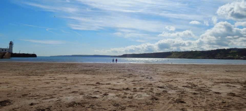 A sandy beach under a partly cloudy sky, with two people walking near the shoreline. A lighthouse stands on a distant pier to the left and hills stretch along the right horizon—an inviting scene from a personal guide to Scarborough.