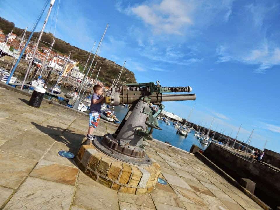 A young boy looks through a large, old naval telescope mounted on a stone platform by a harbour—your personal guide to Scarborough’s charming boats, hillside buildings, and blue skies in the background.