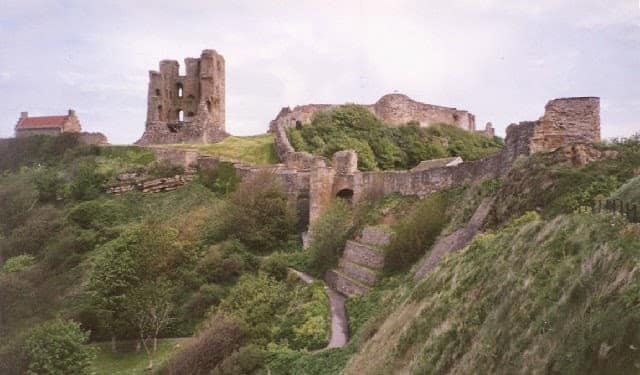 Ruins of an old stone castle sit atop a grassy hill, surrounded by greenery and partially overgrown walls, with a cloudy sky in the background—a scene straight from A personal guide to Scarborough. Steps and pathways wind up towards the ruins.