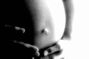 Close-up, black and white image of a pregnant belly, gently cradled by two hands, highlighting the navel. The serene scene evokes tenderness and the intimate moments experienced alongside heartburn during pregnancy. Softly blurred background with bright lighting.