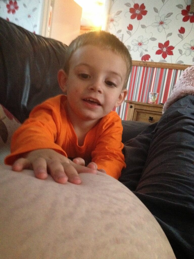 A young boy in an orange shirt touches a pregnant belly with visible stretch-marks while sitting on a sofa. He smiles and looks at the belly, embracing body positivity. The background features floral wallpaper and a wooden cupboard.