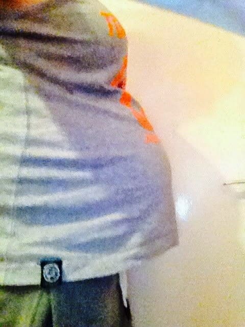 A person in a grey shirt is shown from the side, with a visibly pregnant belly—capturing a Week 27 pregnancy. The background is out of focus and appears to be indoors.