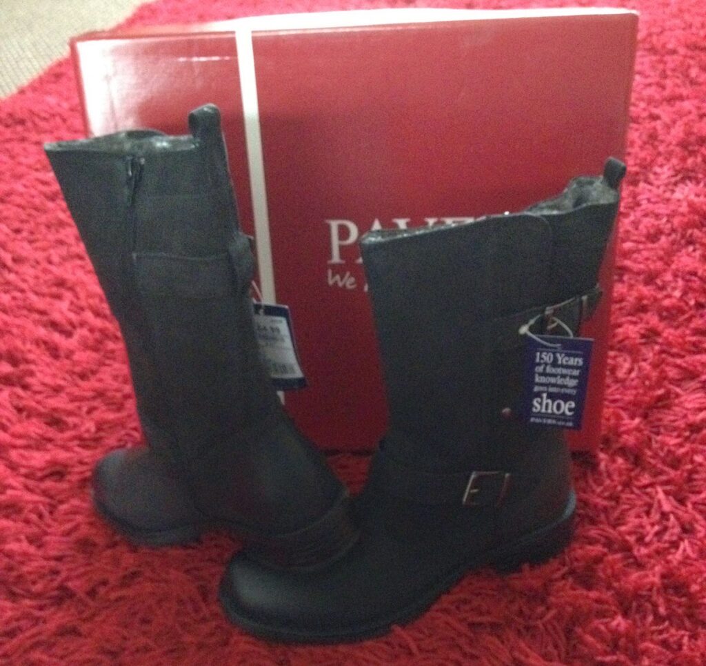 A pair of black Nubuck Leather Calf Boots with buckles and tags is displayed on a red textured rug, with a large red shoebox in the background.