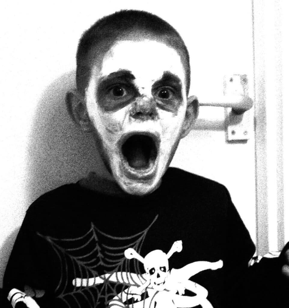 A child in a skeleton costume and dramatic skull face paint opens their mouth wide in a surprised or scared expression, capturing the spooky spirit of Hallowe’en Adventures. The image is in black and white.