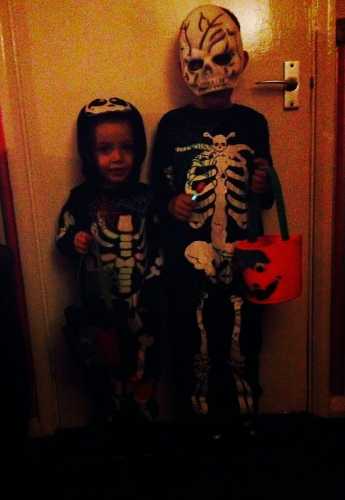 Two children in skeleton costumes, one with a skull mask, stand indoors holding Halloween buckets, ready for spooky adventures. The dim lighting highlights their glow-in-the-dark skeleton designs as they prepare to go trick-or-treating.