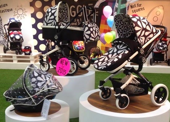 Three modern Cosatto baby pushchairs with bold black and white floral patterns are displayed on round platforms at an indoor press launch, surrounded by balloons, colourful decor, and other vibrant baby things.