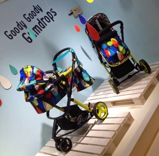 Two colourful Cosatto baby pushchairs with geometric patterns are displayed on white plinths against a blue wall with the words "Goody Goody Gumdrops" and decorative raindrop shapes—perfect for a stylish press launch of new baby things.