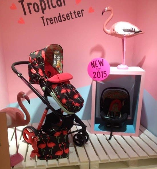 A Cosatto pushchair and car seat with a flamingo pattern are displayed in a pink and blue setting with flamingo decorations. Perfect for press launch photos, these baby items stand out under the sign “Tropical Trendsetter” and "NEW for 2015.