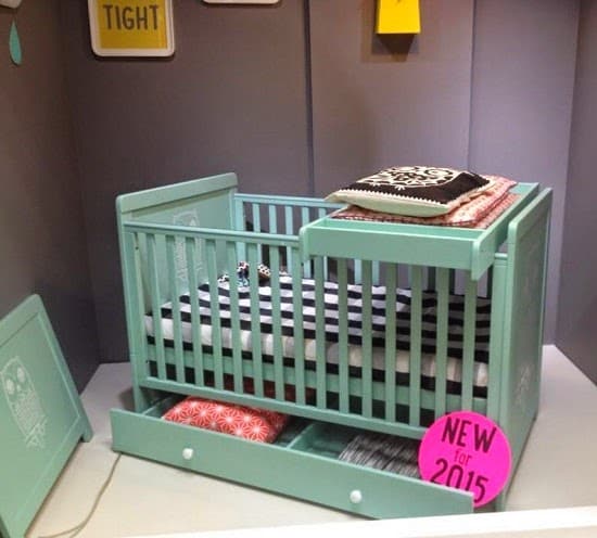 A mint green Cosatto baby cot with pillows inside and on top, featuring a striped mattress, two drawers underneath, and a pink sign that reads "NEW for 2015" on the lower front panel—perfect baby things for any press launch.