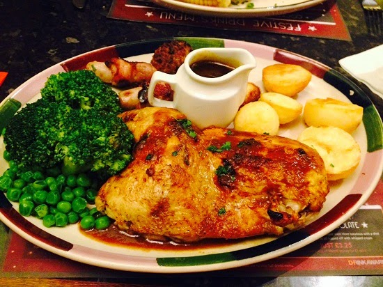 A festive plate from Frankie & Benny's Christmas Menu: roast chicken with gravy, surrounded by roast potatoes, peas, broccoli, pigs in blankets, and a Yorkshire pudding. A small gravy jug sits on the plate.