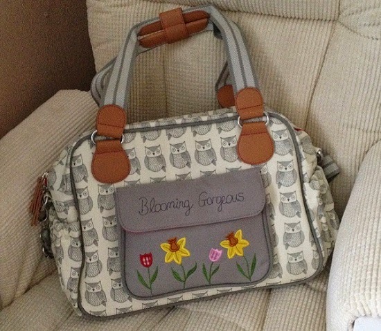 A grey and white Pink Lining changing bag with an owl pattern, brown handles, and a front pocket embroidered with "Blooming Gorgeous" and colourful flowers, rests on a beige cushioned chair.