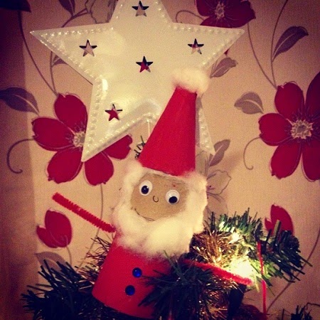 A handmade Father Christmas tree topper with googly eyes, cotton beard, and a red paper hat brings extra cheer to Christmas family time, sitting atop the tree with a white star decoration and red flower wallpaper in the background.