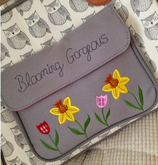A grey Pink Lining changing bag pouch embroidered with "Blooming Gorgeous" and adorned with yellow daffodils plus pink and red tulips, set against an owl pattern background.