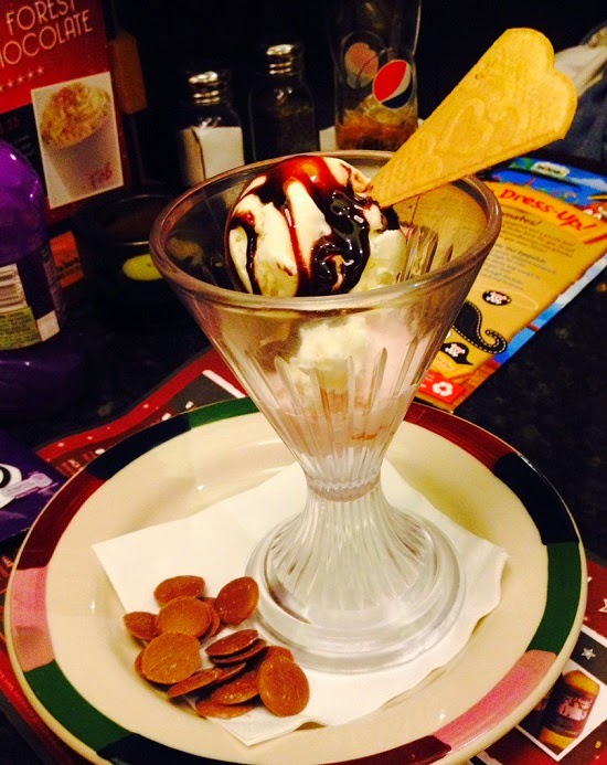 A glass sundae dish with vanilla ice cream, chocolate sauce, and a heart-shaped biscuit, served on a plate with chocolate buttons and a serviette—part of Frankie & Benny's Festive Christmas Menu.