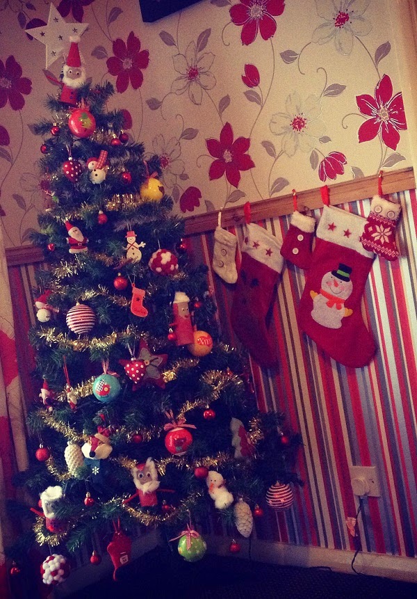 A decorated Christmas tree with tinsel and baubles stands in a festively decorated room, evoking the warmth of Christmas family time. Three stockings hang on a striped wall next to the tree, and floral wallpaper completes the cosy holiday setting.