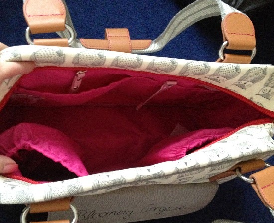 The Blooming Gorgeous changing bag from Pink Lining features a cream colour with charming owl patterns, tan handles, and a pink interior, open to reveal multiple inner pockets and compartments on a blue surface.
