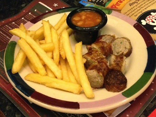 A plate with chips, sliced sausage pieces, and a small black pot of baked beans sits on a table with menus underneath—perfect for enjoying at Frankie & Benny's as part of their festive Christmas Menu.