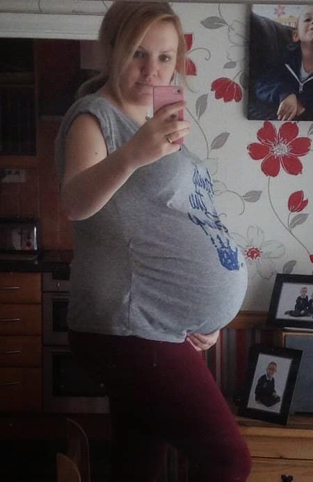 A pregnant woman at week 39 takes a mirror selfie in a cosy home, wearing a grey shirt and holding her bump. Family photos and floral wallpaper can be seen in the background—a sweet pregnancy update.
