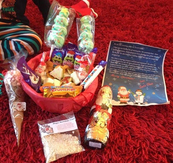 A festive red hamper filled with Christmas-themed sweets, chocolates, and treats sits on a bright red carpet—perfect for sharing with family at Christmas time. Nearby are a chocolate Father Christmas, a letter from Father Christmas, and a bag labelled "Magic Reindeer Food.