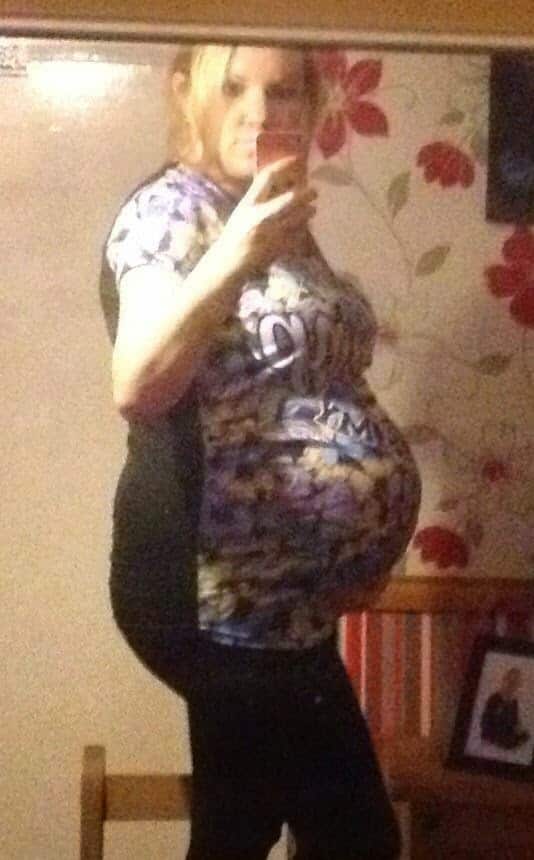 A person wearing a colourful shirt stands sideways in front of a mirror, taking a selfie to share a 36 week pregnancy update. The background features floral wallpaper, a wooden bench, and a framed photo.