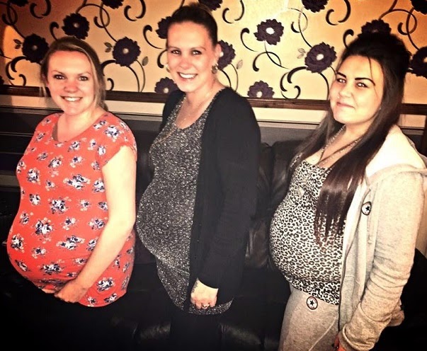 Three smiling pregnant women stand close together, facing the camera. The cosy indoor setting, with a dark sofa and floral wallpaper, captures the warmth of family at Christmas Time.