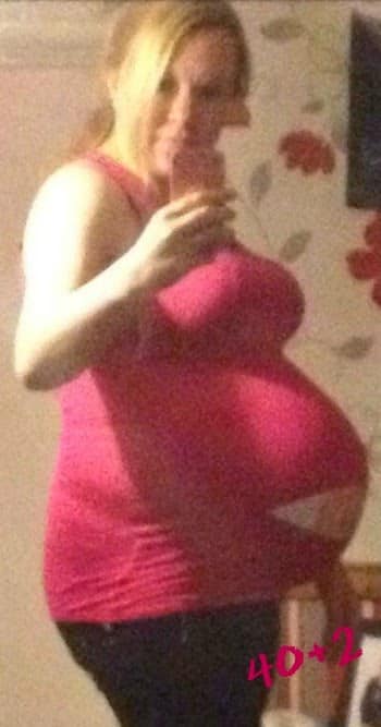 A person wearing a pink vest top and black trousers takes a mirror selfie, proudly showing a heavily pregnant bump. The photo reads "40+2" in pink, marking one week overdue in the waiting game of pregnancy.