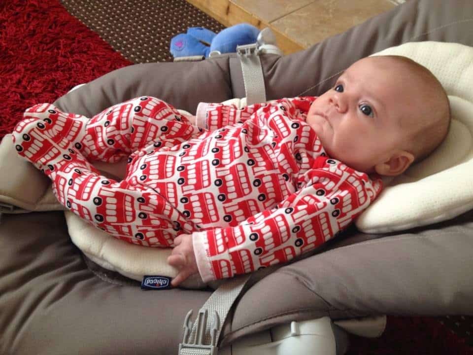 A baby lies in a cushioned baby seat, wearing a red and white babygro patterned with double-decker buses. The baby looks up with a calm expression, captured perfectly for this Week In Pictures. The background features a red carpet and brown rug.