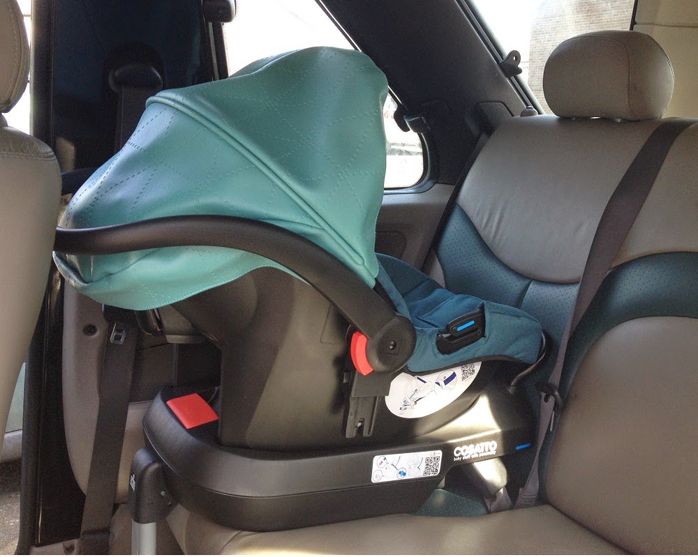 A blue Cosatto Ooba infant car seat is securely installed rear-facing on the back seat of a car, with the carrier’s canopy extended and safety labels visible on the base—perfect for use with this stylish 3-in-1 travel system.