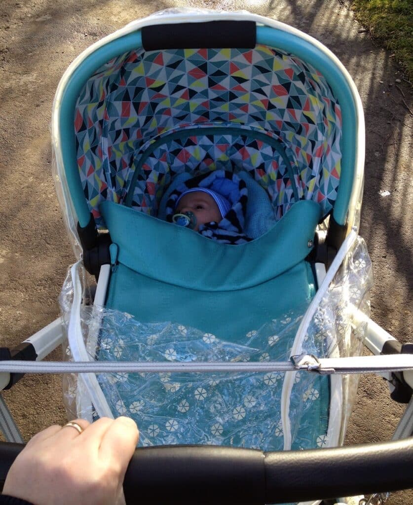 A baby bundled in a blue blanket lies in a Cosatto Ooba pushchair with a colourful, geometric-patterned canopy and clear rain cover. An adult's hand is seen pushing this 3-in-1 Travel System outdoors on a sunny day.