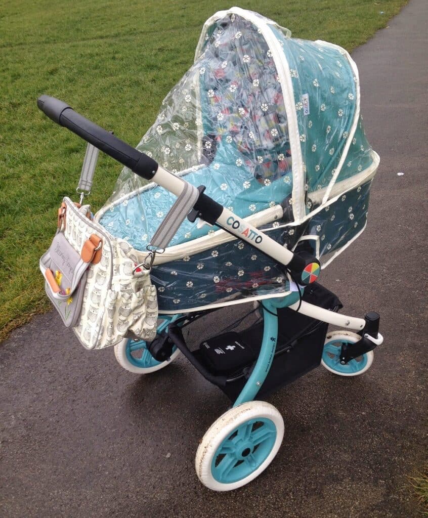 A teal and white Cosatto Ooba 3-in-1 Travel System pushchair with a transparent rain cover is parked on a wet path next to green grass. A patterned nappy bag hangs from the pushchair's handle.