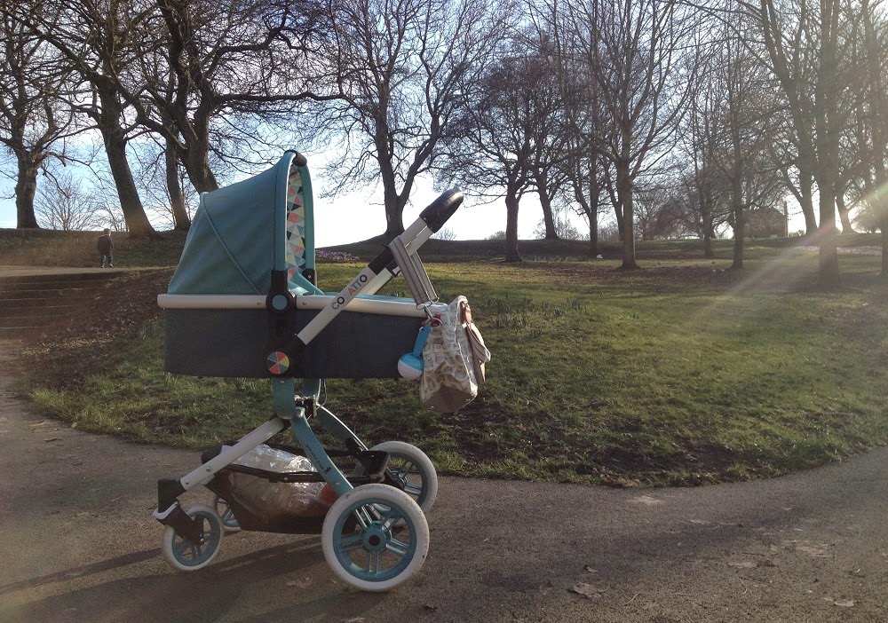 A blue Cosatto Ooba pushchair stands on a paved path in a park with leafless trees and sunlight streaming through the branches. A bag hangs on the pushchair handle, and grass covers the surrounding park area.