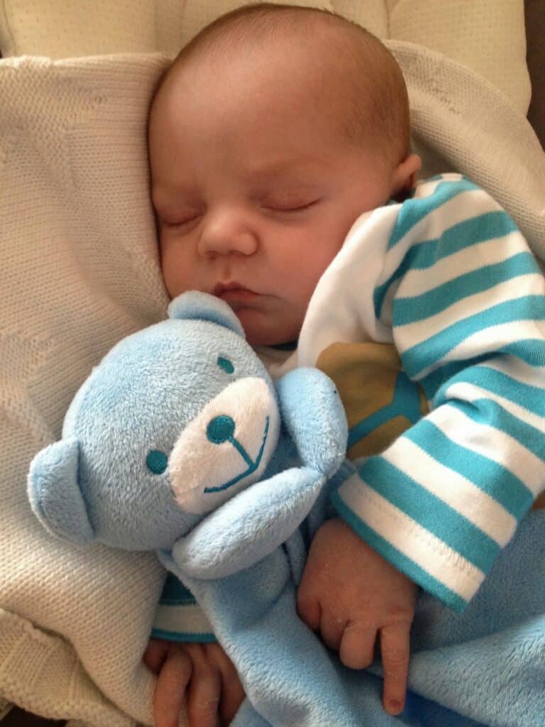 A baby sleeps peacefully whilst cuddling a blue stuffed teddy bear, dressed in a striped blue and white outfit and wrapped in a soft blanket—a sweet moment that warms every parenting journey.