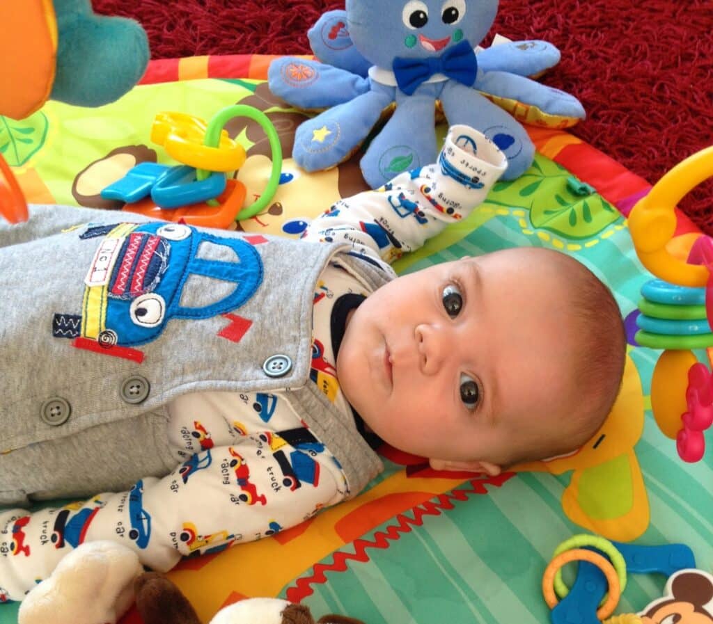 A baby—the youngest child—lies on a colourful play mat surrounded by toys, including a soft octopus and plastic rings. Wearing a patterned baby grow and grey dungarees with a blue car design, the little one looks up at the camera, capturing precious parenting moments.