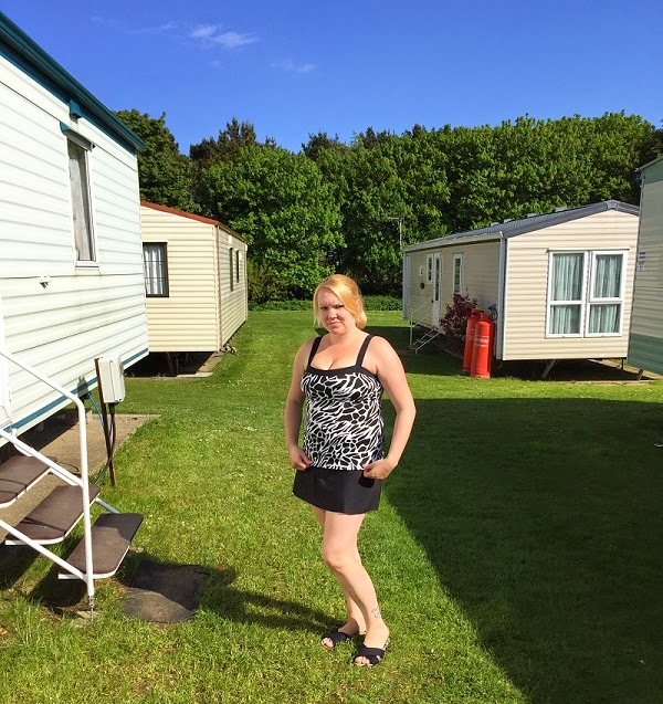 A woman in a black and white Miraclesuit Skirt stands on grass between caravans, with trees and a blue sky in the background.