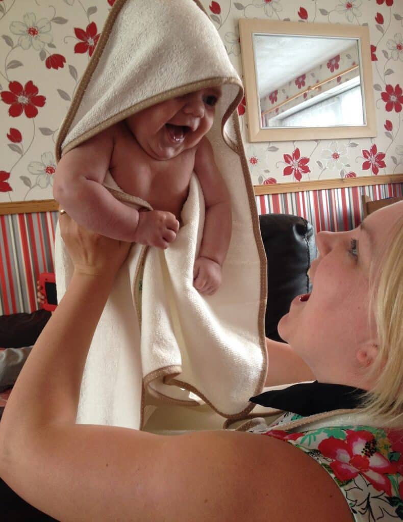 A smiling baby wrapped in a Cuddledry Baby Towel is held up by a woman with blonde hair. Both are joyful, and the room has red floral wallpaper and a mirror reflecting part of the scene.