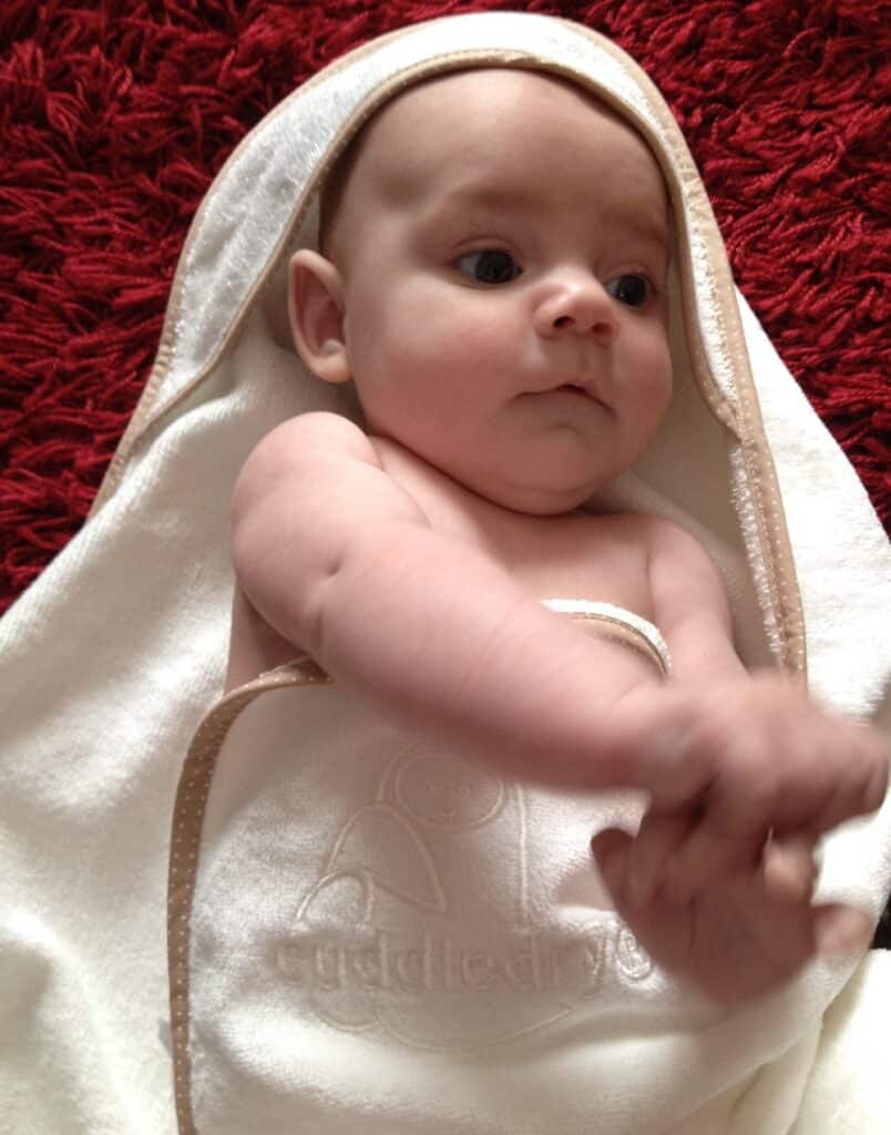 A baby wrapped in a white Cuddledry towel with a hood and gingham edge lies on a red textured surface, gazing upwards with wide eyes and holding its own hands.