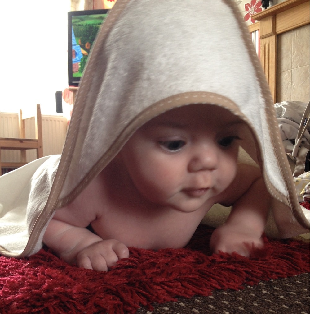 A baby lies on a red carpet, partially covered with a beige Cuddledry baby towel draped over their head, looking forward with wide eyes. A television and furniture are visible in the background.