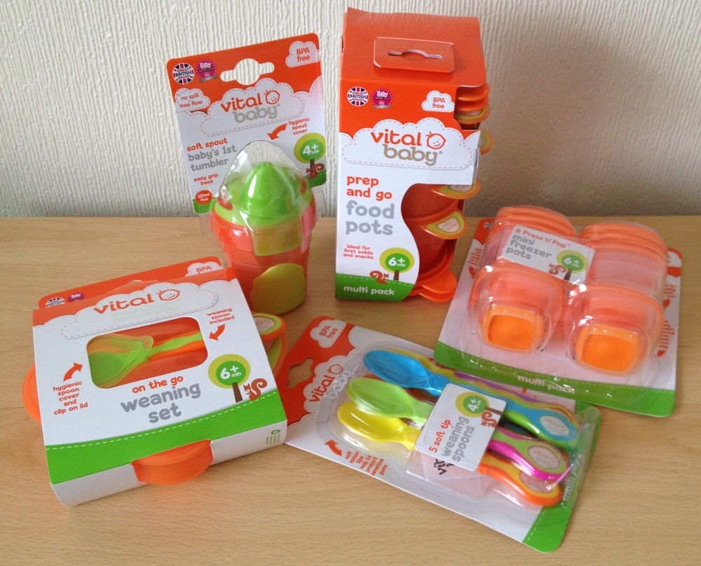 Assorted Vital Baby feeding products in bright packaging, including weaning stage essentials like a weaning set, prep and go food pots, spoons, and a spot and tumble cup, arranged on a wooden surface against a light wall.