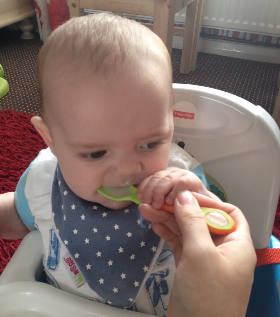 A baby sitting in a highchair is being fed with a spoon by an adult during the weaning stage. The little weaning warrior wears a blue and white bib with stars and holds onto the spoon with one hand.