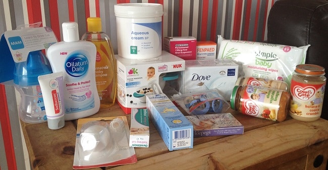 A variety of baby essentials, including lotion, shampoo, baby wipes, a dummy, baby food jars, feeding bottles, cotton wool pads, cream, and soap, are arranged on a wooden table with a striped background.