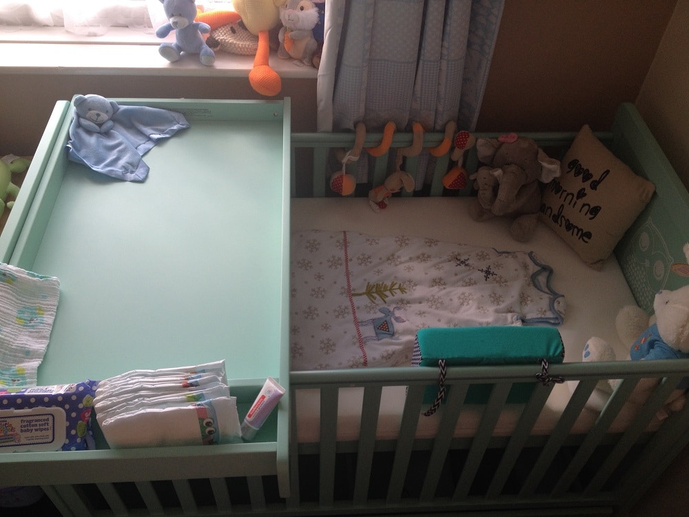 A mint green Cosatto cotbed with an attached changing table, baby clothes, wipes, and toys. The cot features a blanket, soft toys, a “Good morning handsome” cushion, and a mobile with hanging animals above the mattress.