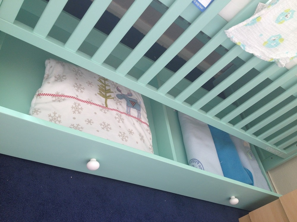 A close-up view of the underside of a mint green Cosatto Story cot bed with an open storage drawer. The drawer contains a folded blanket with a winter design and another folded item, possibly a blanket or towel.