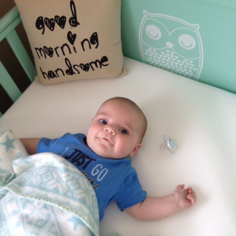A baby lies in a cot, partly covered with a blue patterned blanket. The mint green rail hints at an Owlet Cotbed, with a “good morning handsome” cushion and white owl design on the wall. A dummy rests nearby.