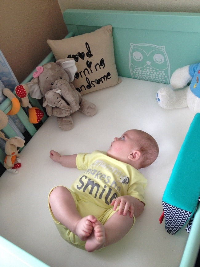A baby in a yellow babygrow lies in an Owlet cot bed, gazing at soft toys. The cot features an owl design and a pillow that says “Good morning, handsome.” The baby is on their back, exploring the surroundings.